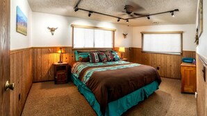 Une chambre à coucher avec un grand lit, des murs en bois, deux fenêtres équipées de stores, un ventilateur de plafond et une petite armoire en bois.