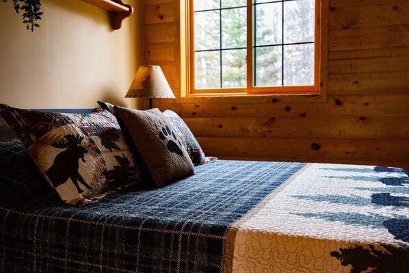 Een slaapkamer in een houten hutje, met een bed, kussens, een raam en een lamp.