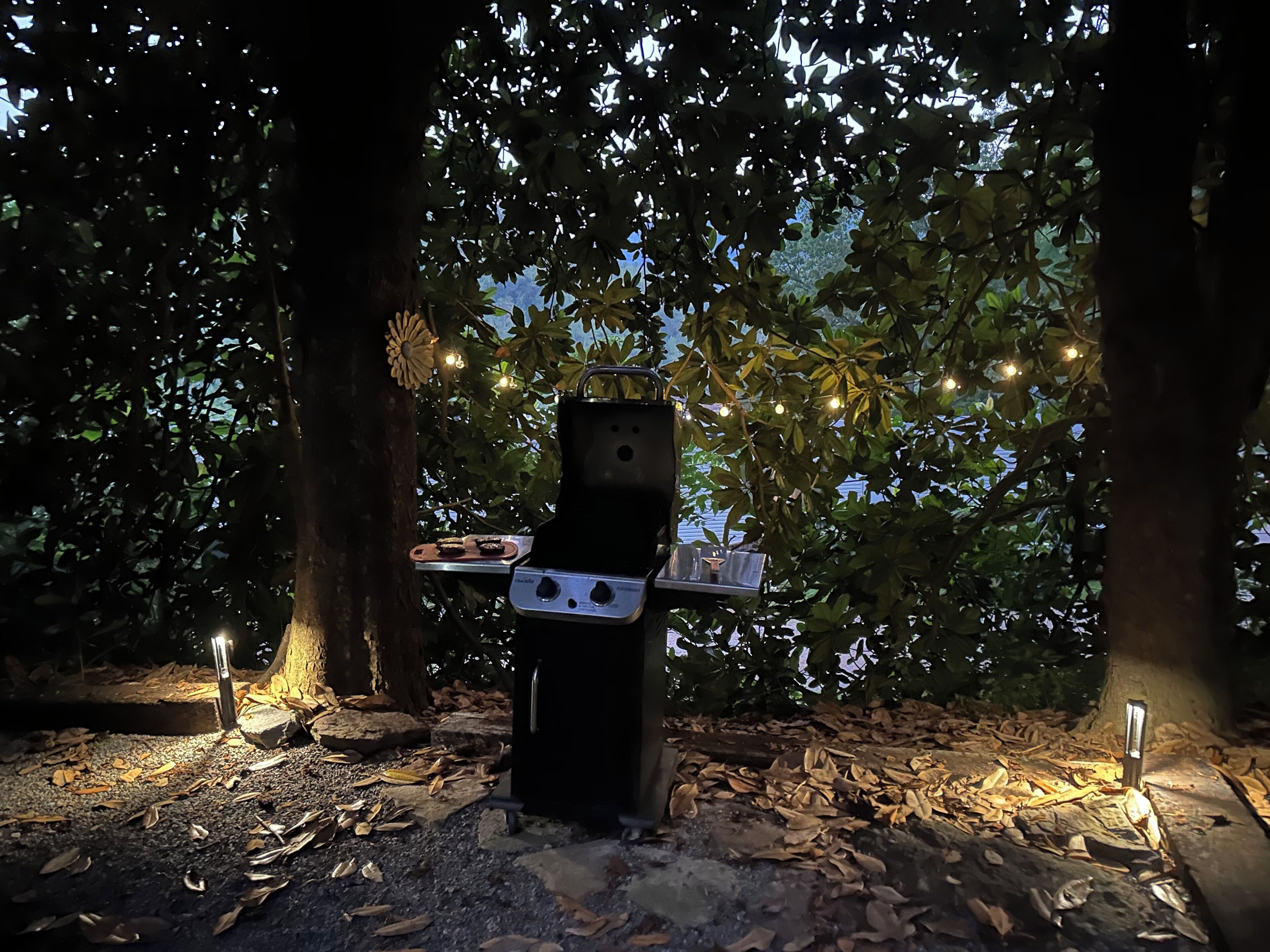 Een buitenkeuken met een tafel en stoelen, omringd door bomen en lichtjes.