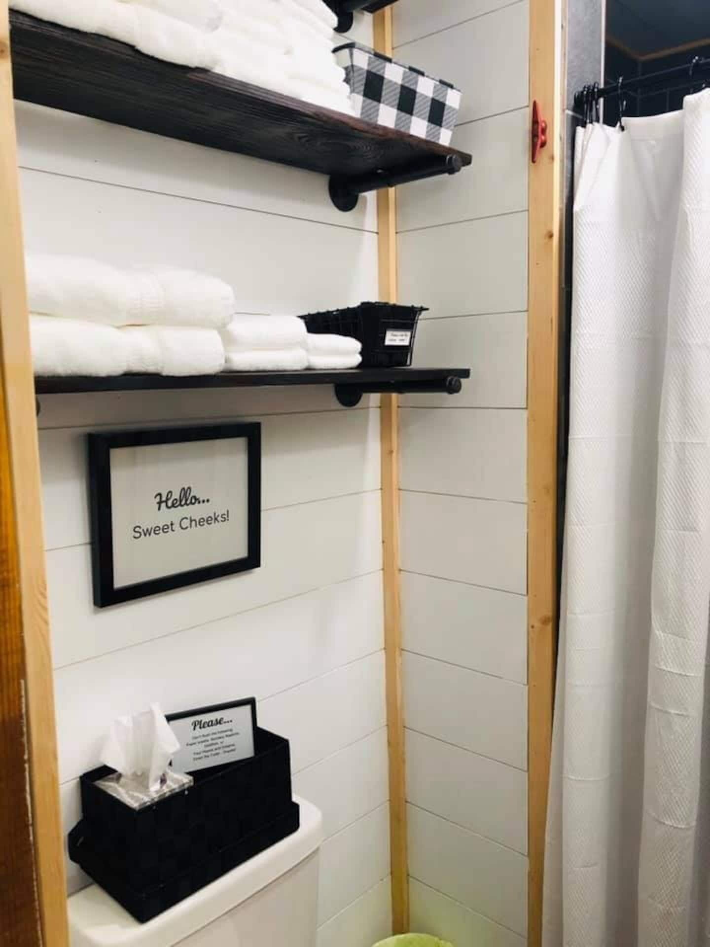 Une salle de bain avec des murs recouverts de carreaux blancs, une porte en bois, un porte-serviettes noir, un panneau encadré sur lequel on peut lire « Bonjour… jolies fesses ! », et une poubelle noire.
