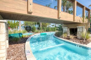 A swimming pool with a wooden bridge overhead, surrounded by greenery and a clear sky.