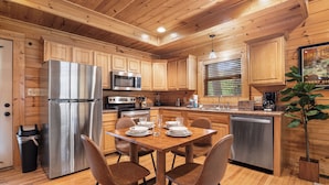 A modern kitchen with wooden cabinets, stainless steel appliances, and a dining area with four chairs. - Mtn Cabin with Hot Tub, pool table, arcade, smartTVs, fast FiWi, great location! (Pigeon Forge)