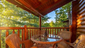 A wooden deck with wicker chairs and a glass table, surrounded by trees. - Mtn Cabin with Hot Tub, pool table, arcade, smartTVs, fast FiWi, great location! (Pigeon Forge)