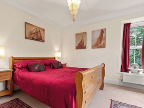 Une chambre à coucher avec un lit en bois, une literie rouge, deux tableaux représentant des voiliers accrochés au mur, une fenêtre avec des rideaux rouges et une table de chevet avec une lampe.