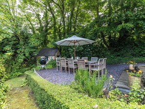 Une terrasse extérieure avec une table et des chaises, entourée d’une végétation luxuriante et d’un petit bâtiment en pierre.