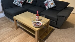 Un salón con un sofá gris oscuro, dos cojines estampados, una mesa de centro de madera con un bol de bombones, una botella de vino tinto y dos copas de vino.