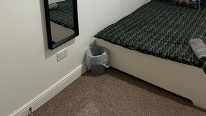 Un dormitorio con una cama individual, un espejo grande, una ventana con persianas, un cesto de basura y suelo alfombrado.