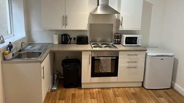 Una cocina compacta con armarios blancos, una campana extractora de acero inoxidable, un microondas, un tostador y un refrigerador pequeño.