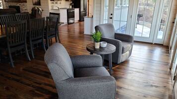 Un salón amplio con piso de madera, sillones grises, una pequeña mesa redonda con una planta y una cocina al fondo.