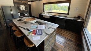 Una cocina con encimera de mármol, taburetes de madera, un refrigerador, un reloj y una ventana con vistas a la vegetación.