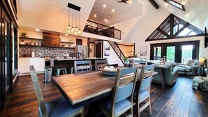 A spacious loft-style living area with a wooden dining table, blue cushioned chairs, a staircase, and a kitchen with a black and white backsplash. - Lakeside | Hot Tub | Kayaks | Fishing (MENA)