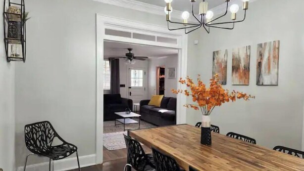 Un coin repas avec une table en bois, des chaises noires et un lustre. Les murs sont ornés de peintures abstraites et un vase contenant des fleurs oranges y est posé.