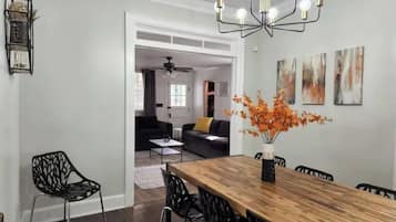 Un coin repas avec une table en bois, des chaises noires et un lustre. Les murs sont ornés de peintures abstraites et un vase contenant des fleurs oranges y est posé.