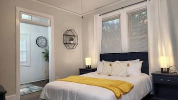 Un dormitorio con cama, dos mesitas de noche, una mesilla, ventilador de techo, reloj y ventana con persianas.