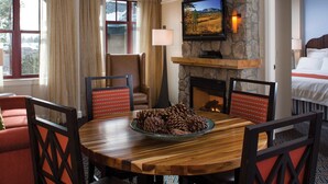 Habitación de hotel con chimenea de piedra, un televisor montado encima, una mesa de comedor con sillas y una cama con manta roja.