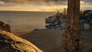 Escena costera con una estructura de piedra, una playa rocosa y una persona de pie sobre una roca.