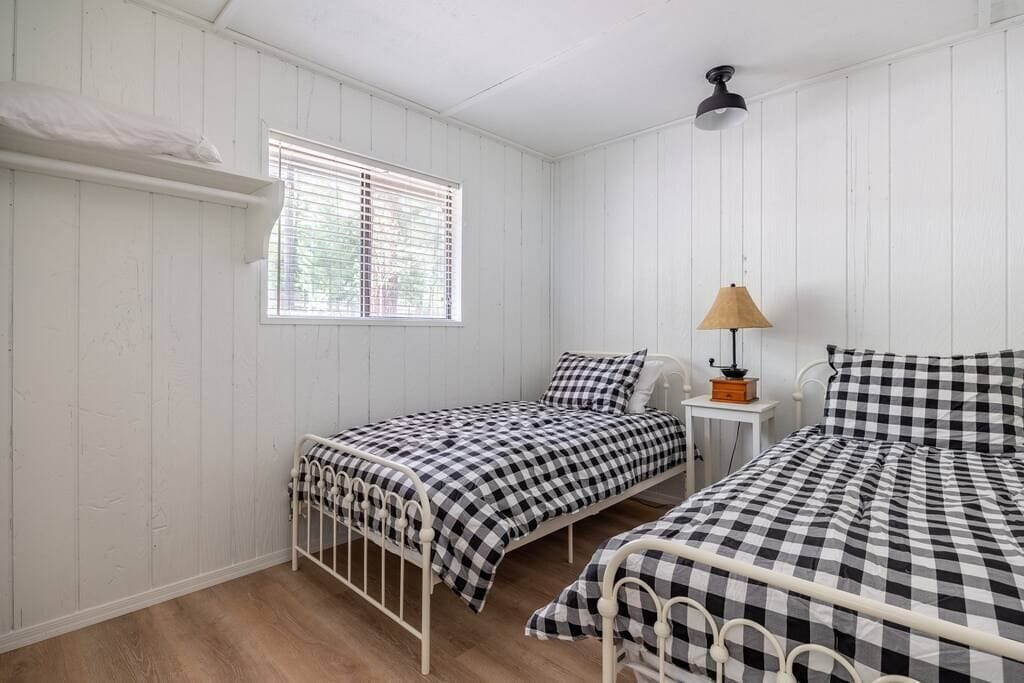 Une petite pièce aux murs recouverts de panneaux blancs, avec deux lits simples, une fenêtre avec des stores, une table de chevet avec une lampe et une étagère au-dessus de la fenêtre.