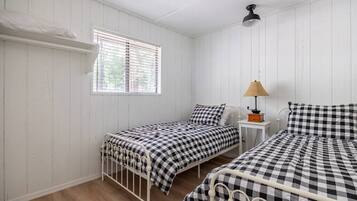 Une petite pièce aux murs recouverts de panneaux blancs, avec deux lits simples, une fenêtre avec des stores, une table de chevet avec une lampe et une étagère au-dessus de la fenêtre.