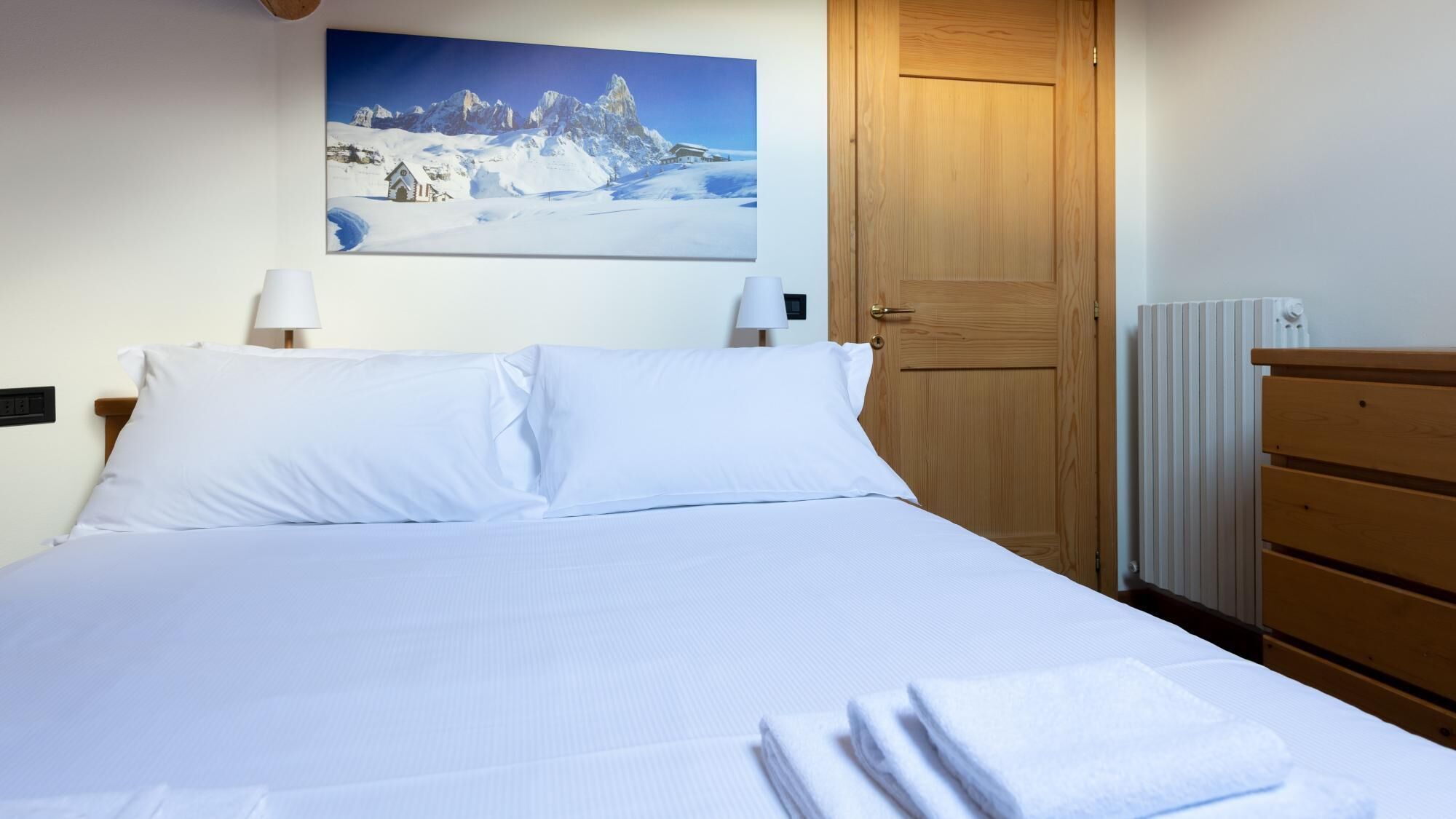 A neatly made bed with white linens, a wooden dresser, a door, and a painting of a snowy mountain landscape on the wall.