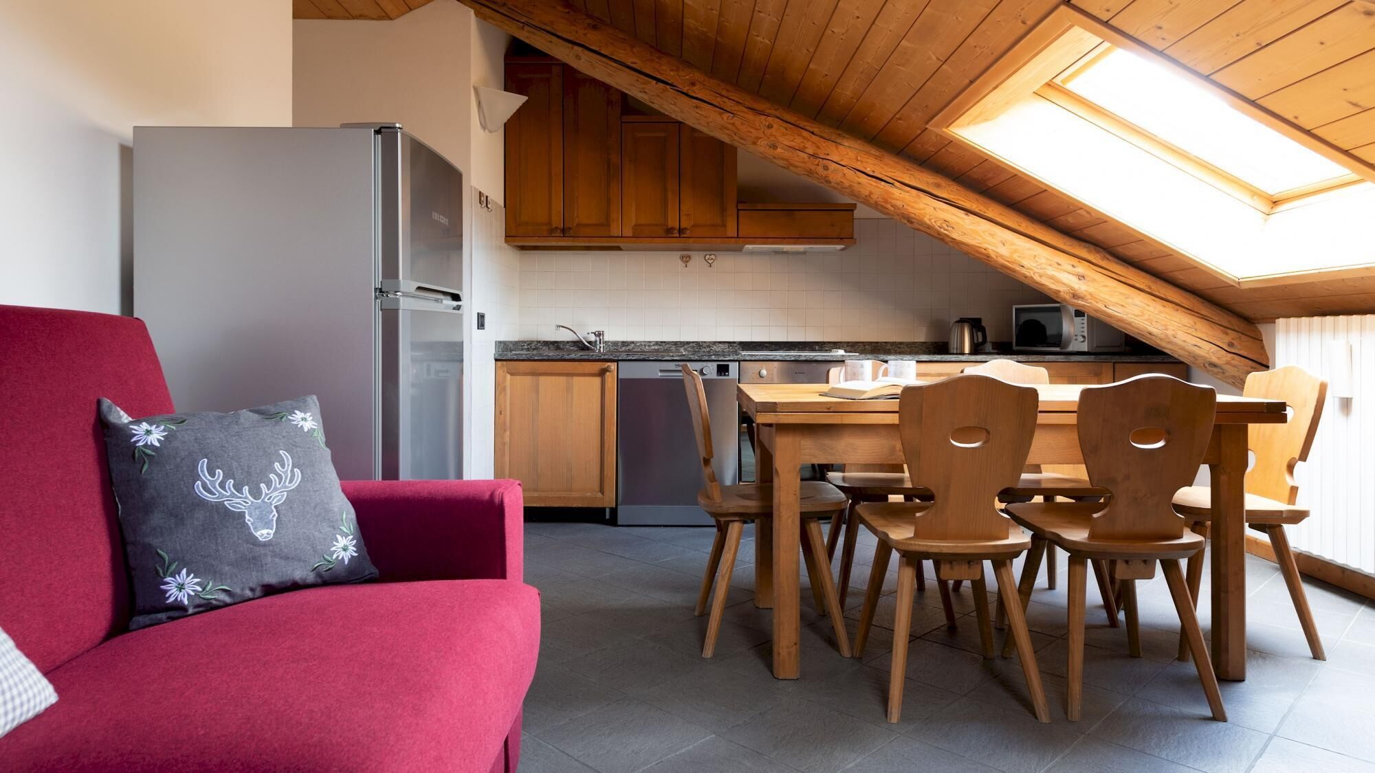 A cozy kitchen with wooden cabinets, a dining table with chairs, a red sofa with a decorative pillow, and a skylight above.