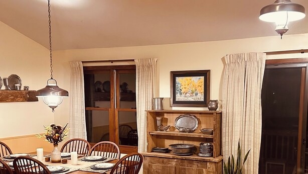 A dining area with a wooden table, chairs, and hanging pendant lights. -  Cozy Cabin with river frontage yards from river launch for kayaking or fishing. (Cherokee Village)
