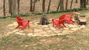 A circular outdoor seating area with red chairs and a fire pit, surrounded by a stone patio, with a lake and trees in the background. -  Cozy Cabin with river frontage yards from river launch for kayaking or fishing. (Cherokee Village)