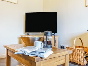 Une table en bois sur laquelle se trouve un livre ouvert, une cafetière et une tasse. Un téléviseur est fixé au mur derrière la table.