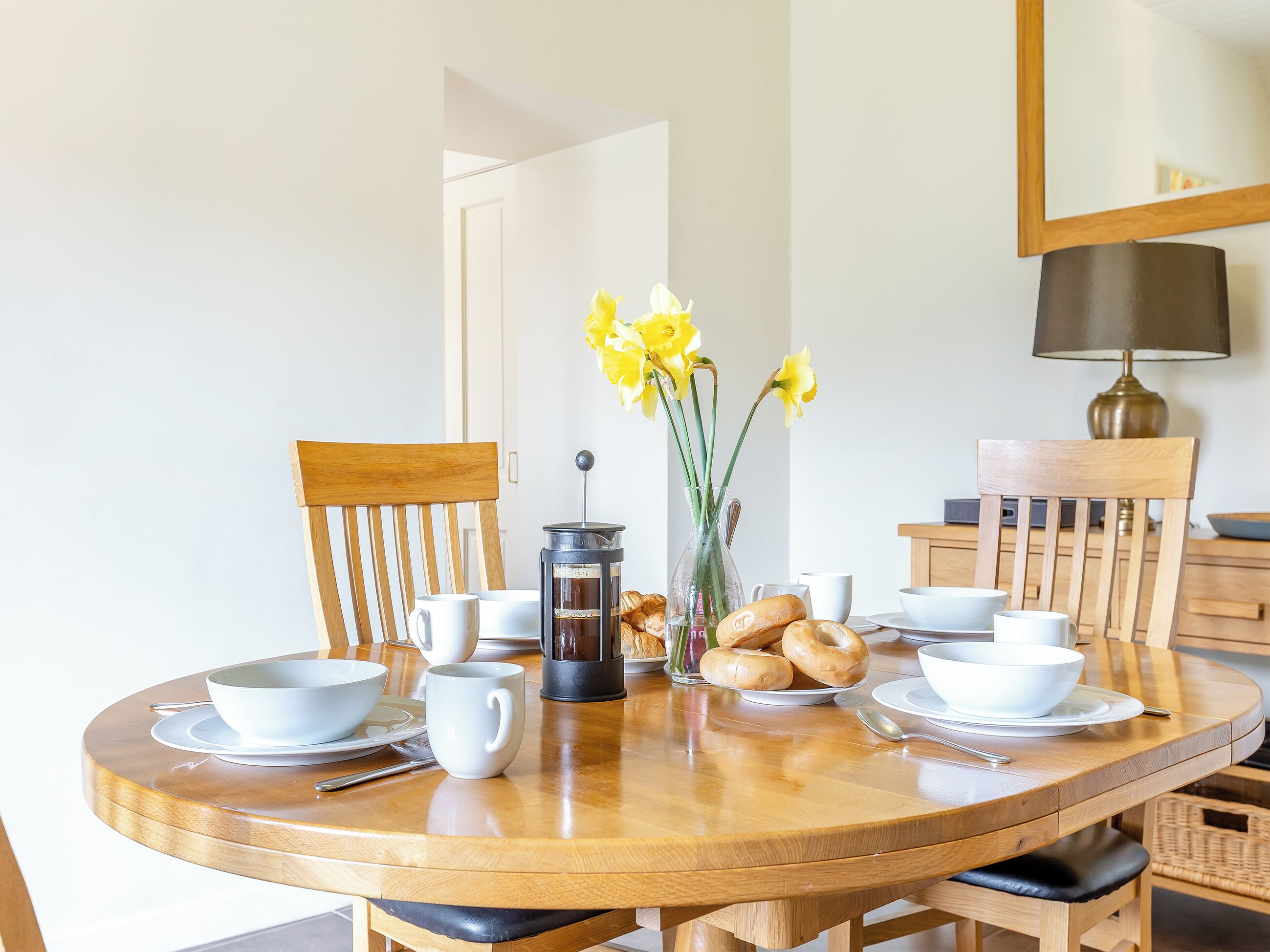 A round wooden dining table with white cups, saucers, a coffee pot, and bread.