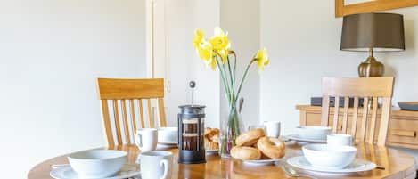 A round wooden dining table with white cups, saucers, a coffee pot, and bread.