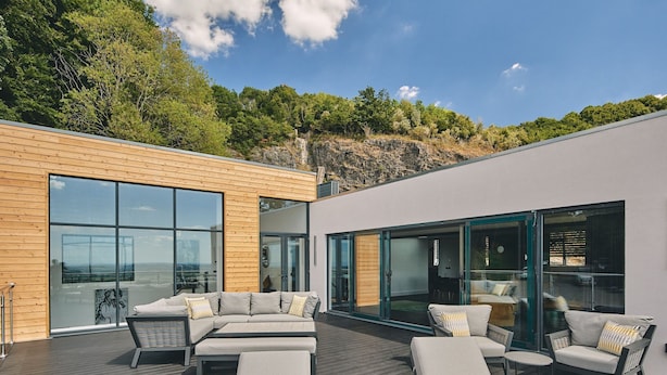 A modern outdoor living area with a wooden deck, large glass windows, and outdoor furniture. - Reservoir View -  a modern home that sleeps 8 guests  in 4 bedrooms (Cheddar)