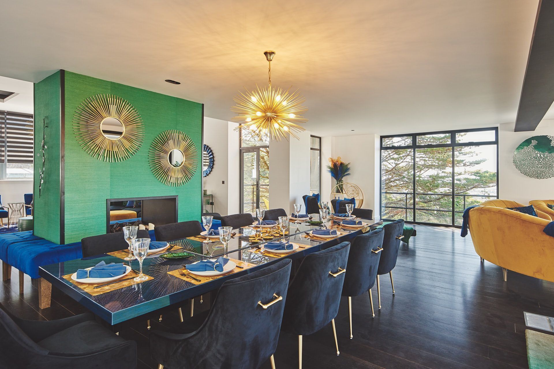 Un espace de salle à manger moderne, avec une longue table dressée pour six personnes, entourée de chaises bleu foncé aux pieds dorés.