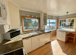 A modern kitchen with wooden cabinets, a granite countertop, and a large window offering a view of a lake and trees. - "Farm House" on Lake of Bays-Muskoka (Waterfront) (Huntsville)