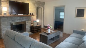 Ein Wohnzimmer mit Kamin aus Stein, einem Flachbildfernseher, grauen Sofas und einem Couchtisch mit Blumenvase.