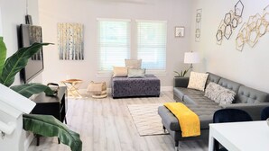 Una sala de estar moderna con un sofá gris, una mesita de centro y un televisor. Hay dos ventanas con persianas, una planta y decoración en la pared.