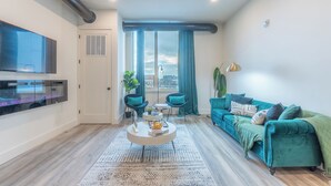 A modern living room with a flat-screen TV, a round coffee table, teal sofas, and a large window with curtains. - Emerald Escape with Free Parking & 2 baths. 5 beds+Sleeps 8!  ✨20% OFF 7 days✨ (East Orange)