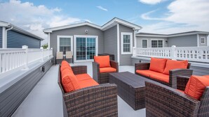 Une terrasse extérieure couverte, aménagée avec des meubles en osier et des coussins orange, un bâtiment gris avec une clôture blanche, et un ciel bleu dégagé.