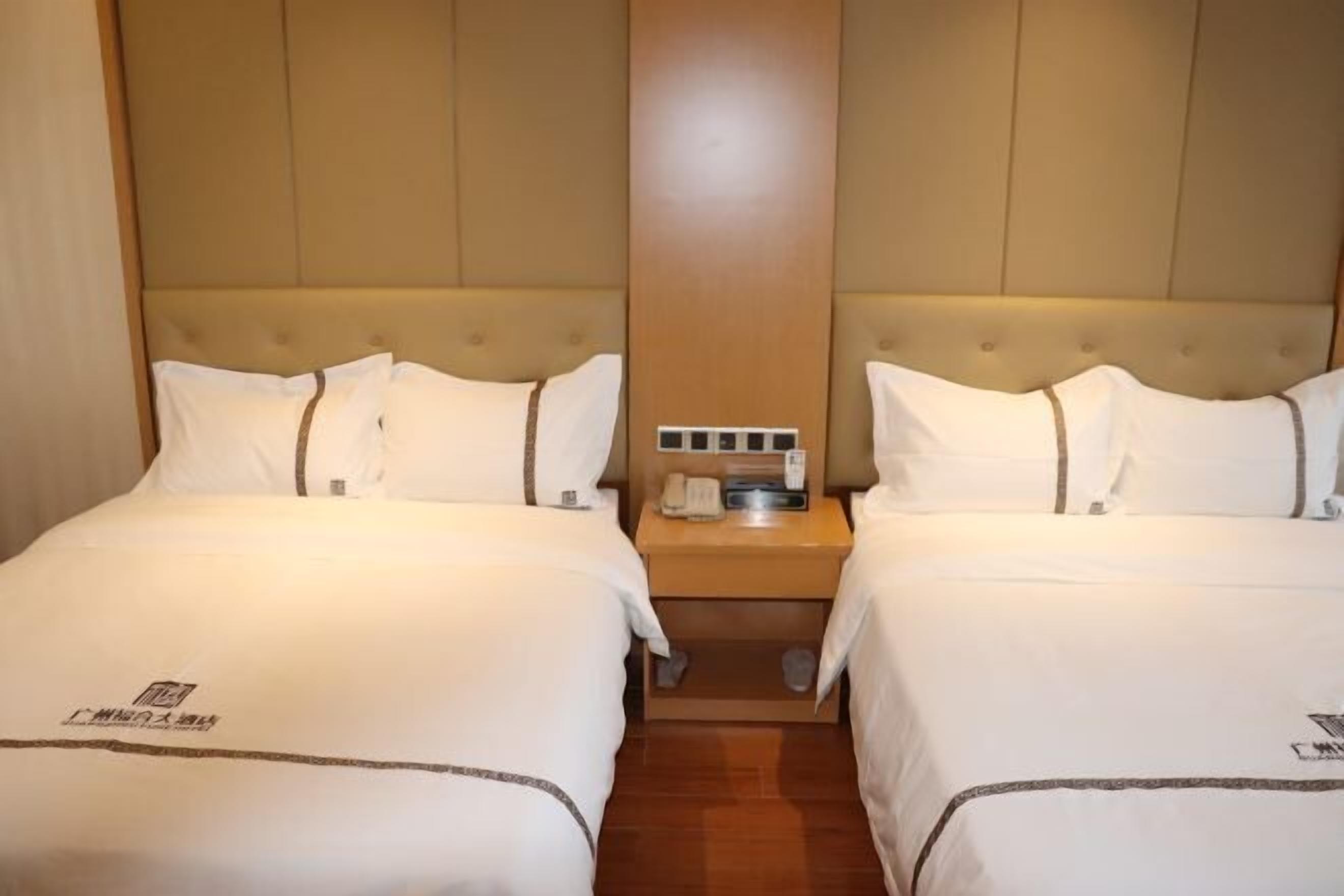 A hotel room with two beds, a wooden headboard, a nightstand with a phone and a lamp, and a small table between the beds.