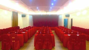 A conference room with rows of tables and chairs, a stage with a curtain, and a door on the right.