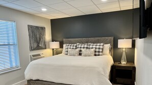 A modern bedroom with a large bed, two bedside tables with lamps, a wall-mounted TV, and a window with blinds.