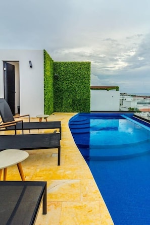 Un área de piscina en la azotea con piscina curva, zona de estar con mesa y sillas, y un muro de seto verde.