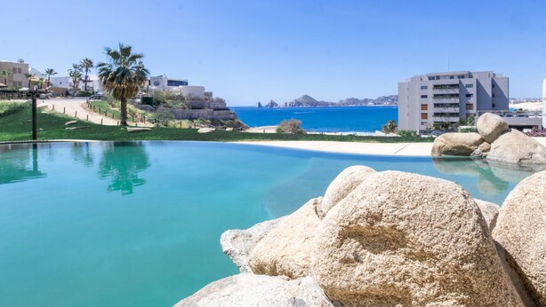 A pool with clear blue water, surrounded by large rocks and palm trees, with a view of the sea and buildings in the background. - Stunning Pool & Ocean Views! Ground Floor Unit! (Cabo San Lucas)