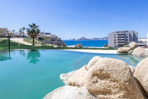 Ein Pool mit klarem, blauem Wasser, umgeben von großen Felsen und Palmen, mit Blick auf das Meer und Gebäude im Hintergrund.