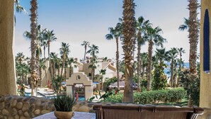 A patio area with a wicker chair and a small table, overlooking a pool and palm trees. - Stunning Pool & Ocean Views! Ground Floor Unit! (Cabo San Lucas)