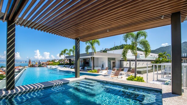 Uma área de piscina moderna com água azul cristalina, espreguiçadeiras e vista para um horizonte urbano com edifícios e uma colina ao fundo.