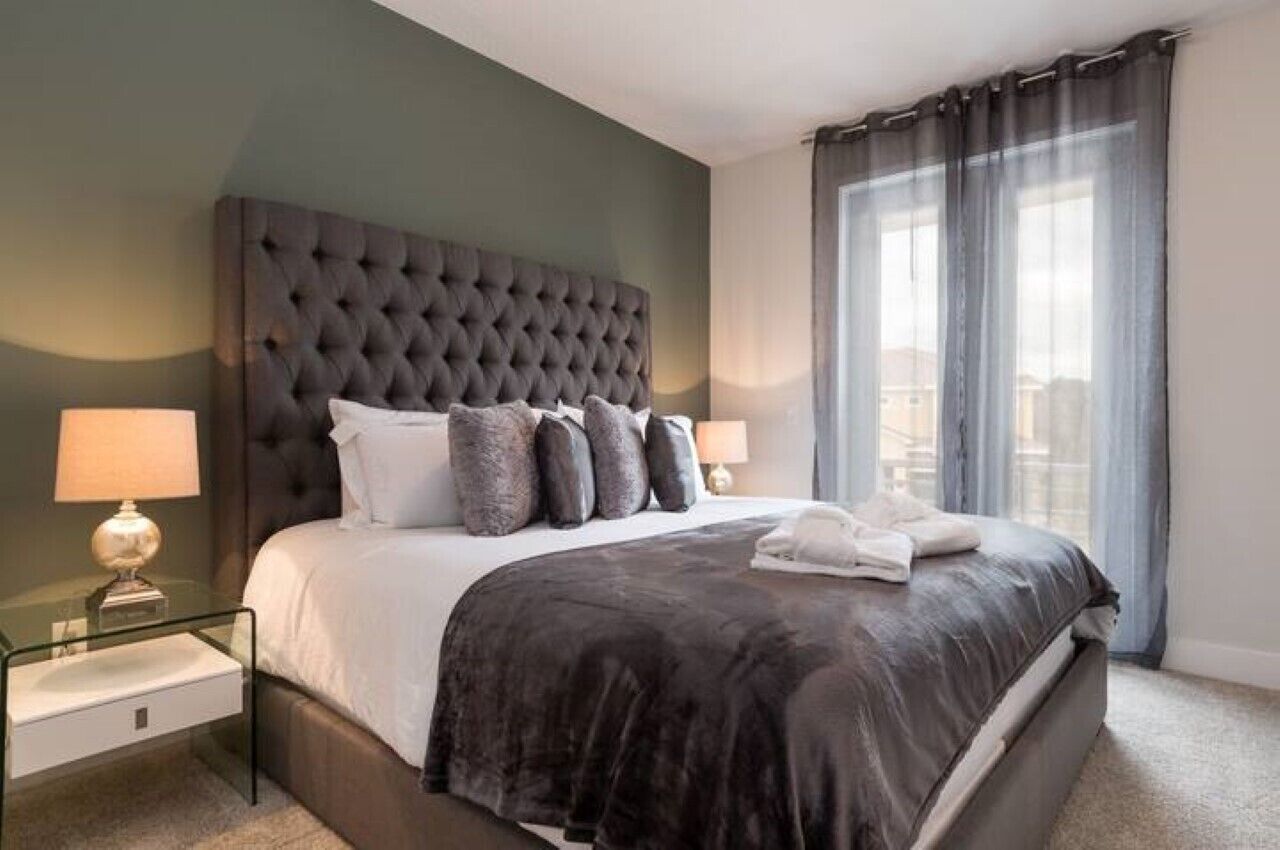 A modern hotel room with a tufted headboard, a large bed with a dark comforter, two bedside lamps, and a view of the city through sheer curtains.