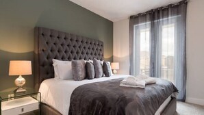 Une chambre d’hôtel moderne avec une tête de lit capitonnée, un grand lit recouvert d’une couette sombre, deux lampes de chevet et une vue sur la ville à travers des rideaux transparents.
