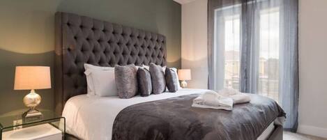 A modern hotel room with a tufted headboard, a large bed with a dark comforter, two bedside lamps, and a view of the city through sheer curtains.