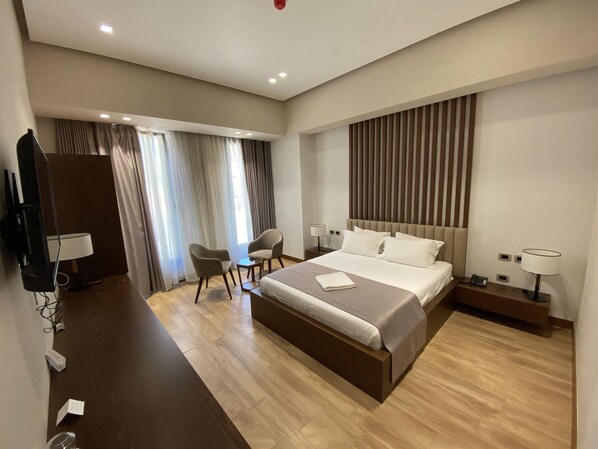 Una habitación de hotel moderna con una cama grande, dos mesitas de noche, un escritorio con una silla, un televisor y un ventanal con cortinas.
