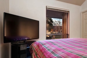 Una habitació amb una gran televisió plana penjada a la paret, un llit amb llençols de colors i ratlles, i una finestra que ofereix vistes a un pati amb tanca de fusta i arbres.