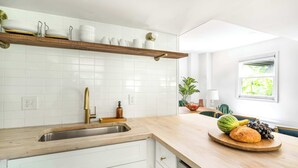 Una cucina con mobili bianchi, un piano di lavoro in legno, una dispensa con stoviglie, un lavello e una finestra vista verde.